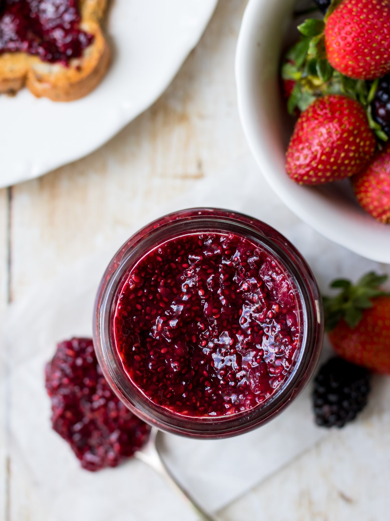 Top down view image of a mixed berry chia jam in jar with a spoon on the side, fresh berries scattered around.