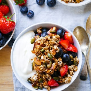 Toasted Muesli made with puffed buckwheat in a small pale blue bowl with yoghurt ahd berries. Extra berries and silver spoons to the side.