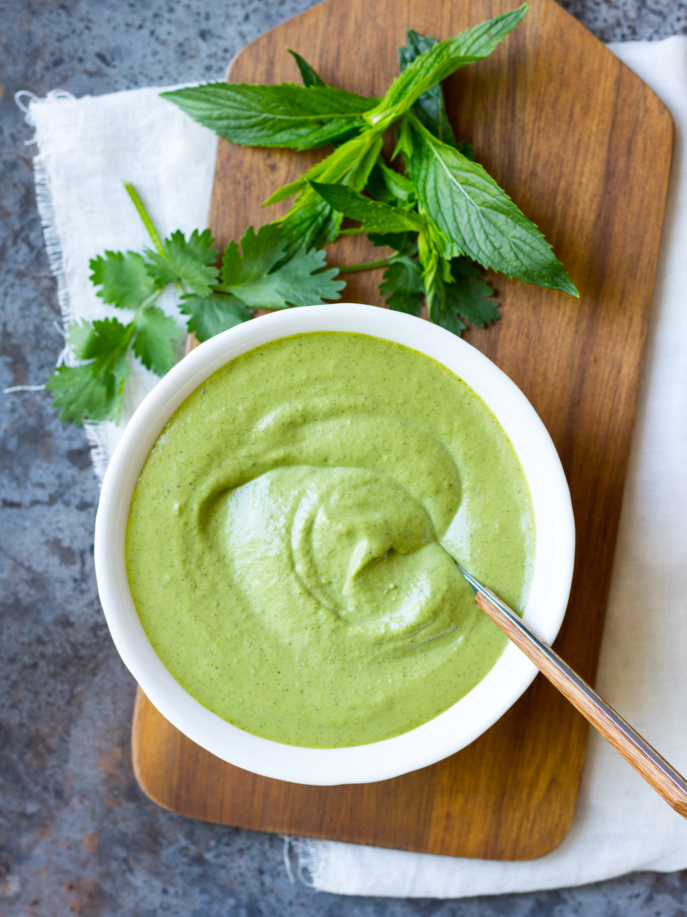 Cashew Tahini Herb Sauce in a white bowl with a spoon, on a chopping board.