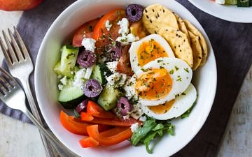 Meal prep breakfast bowls Greek style, combining boiled eggs with chopped cucumber, tomatoes, capsicum, feta and olives.