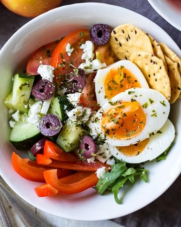 Meal prep breakfast bowls Greek style, combining boiled eggs with chopped cucumber, tomatoes, capsicum, feta and olives.
