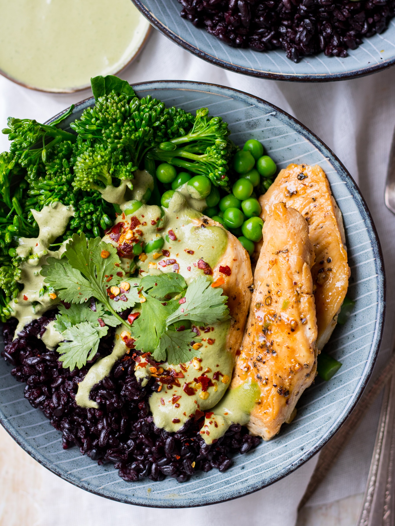 Close up shot of chicken black rice bowls; one blue bowl with black rice, marinated chicken, broccolini, peas, green cashew herb sauce
