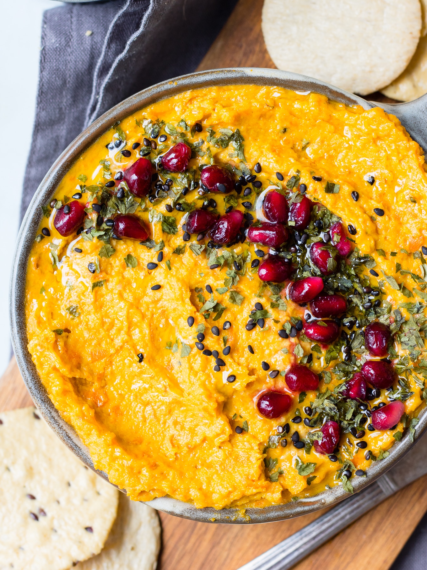 Roasted carrot tahini dip in grey serving bowl topped with sesame seeds and pomegranate and herbs, on chopping board.