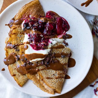 Sweet vegan buckwheat crepes folded on white plate served with yoghurt, berries and chocolate sauce.