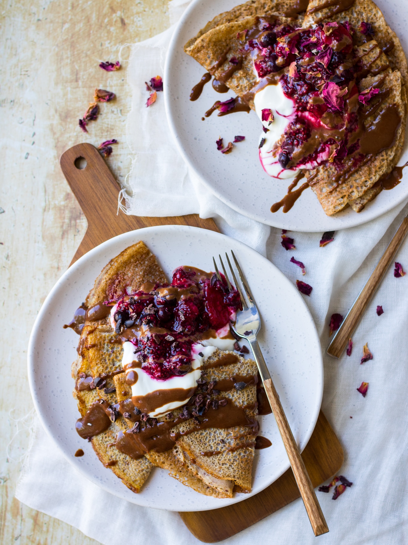 Vegan buckwheat crepes served on a white plate, folded and topped with yoghurt, berry sauce and chocolate sauce drizzle.