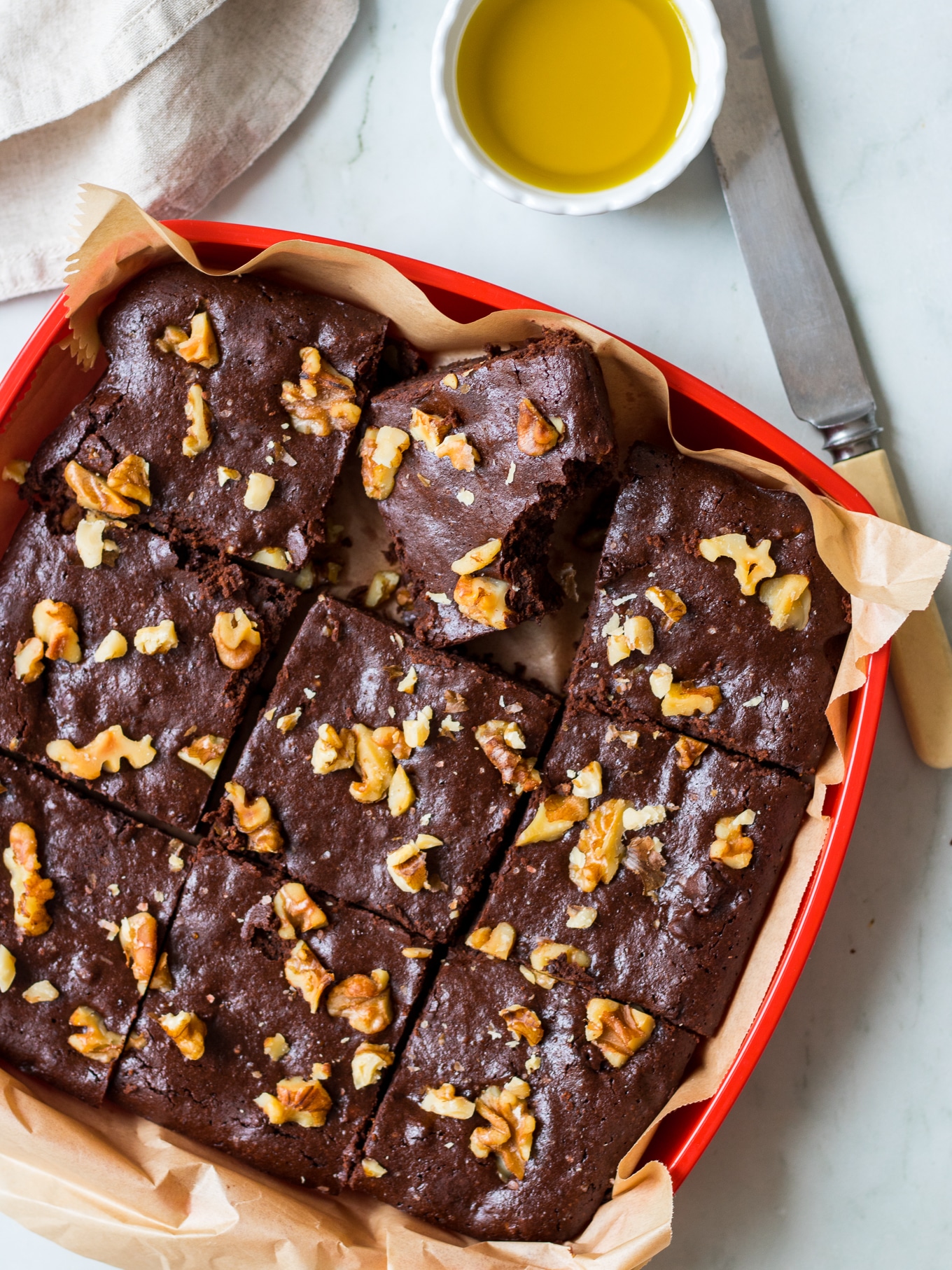 Olive oil chocolate brownies tucked into a red container, on an angle, with butter knife, and olive oil in small dish