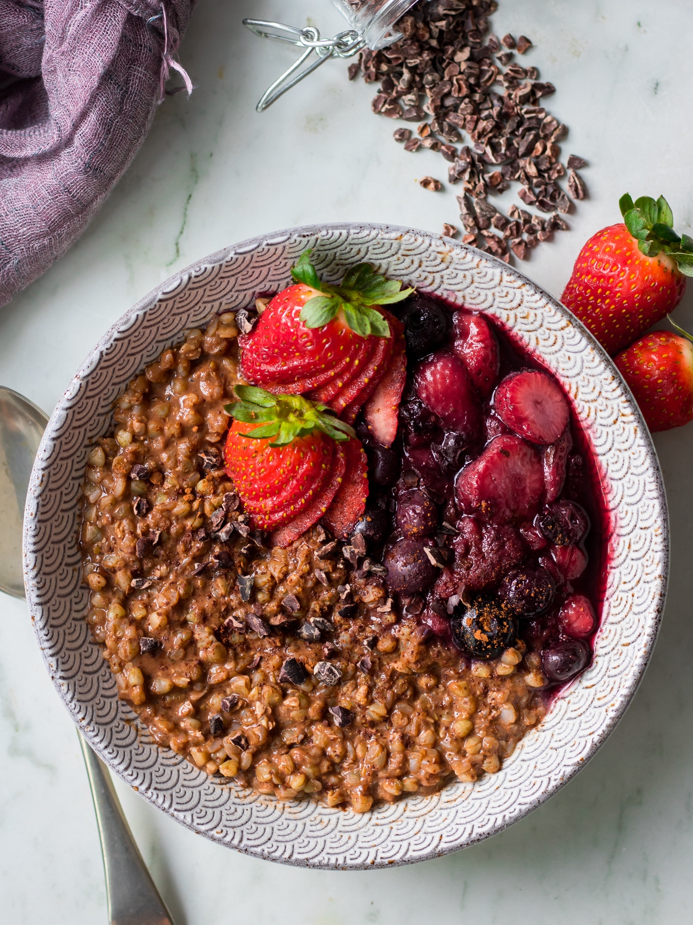 Top view of chocolate buckwheat porridge and stewed berries in patterned bowl, cacao nibs scattered