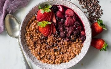Chocolate buckwheat porridge in patterned bowl on marble background, spoon, berries, cacao nibs
