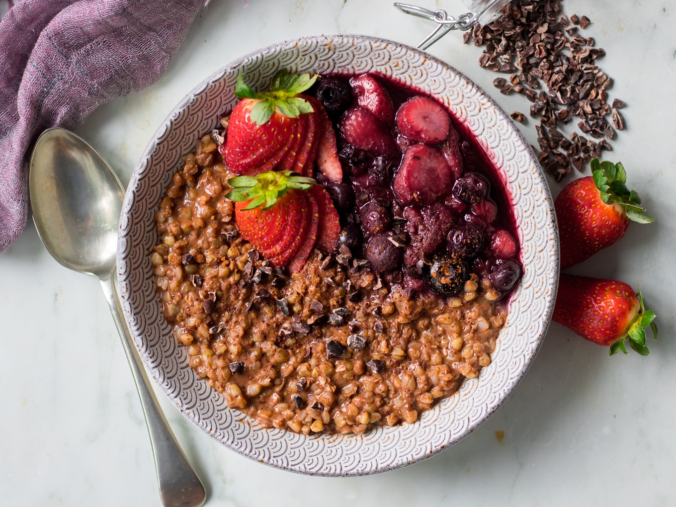 Chocolate buckwheat porridge in patterned bowl on marble background, spoon, berries, cacao nibs