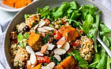 Honey roast pumpkin quinoa salad in grey bowl with rocket, flaked almonds on top, roast pumpkin in background