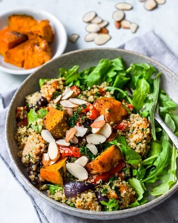 Honey roast pumpkin quinoa salad in grey bowl with rocket, flaked almonds on top, roast pumpkin in background