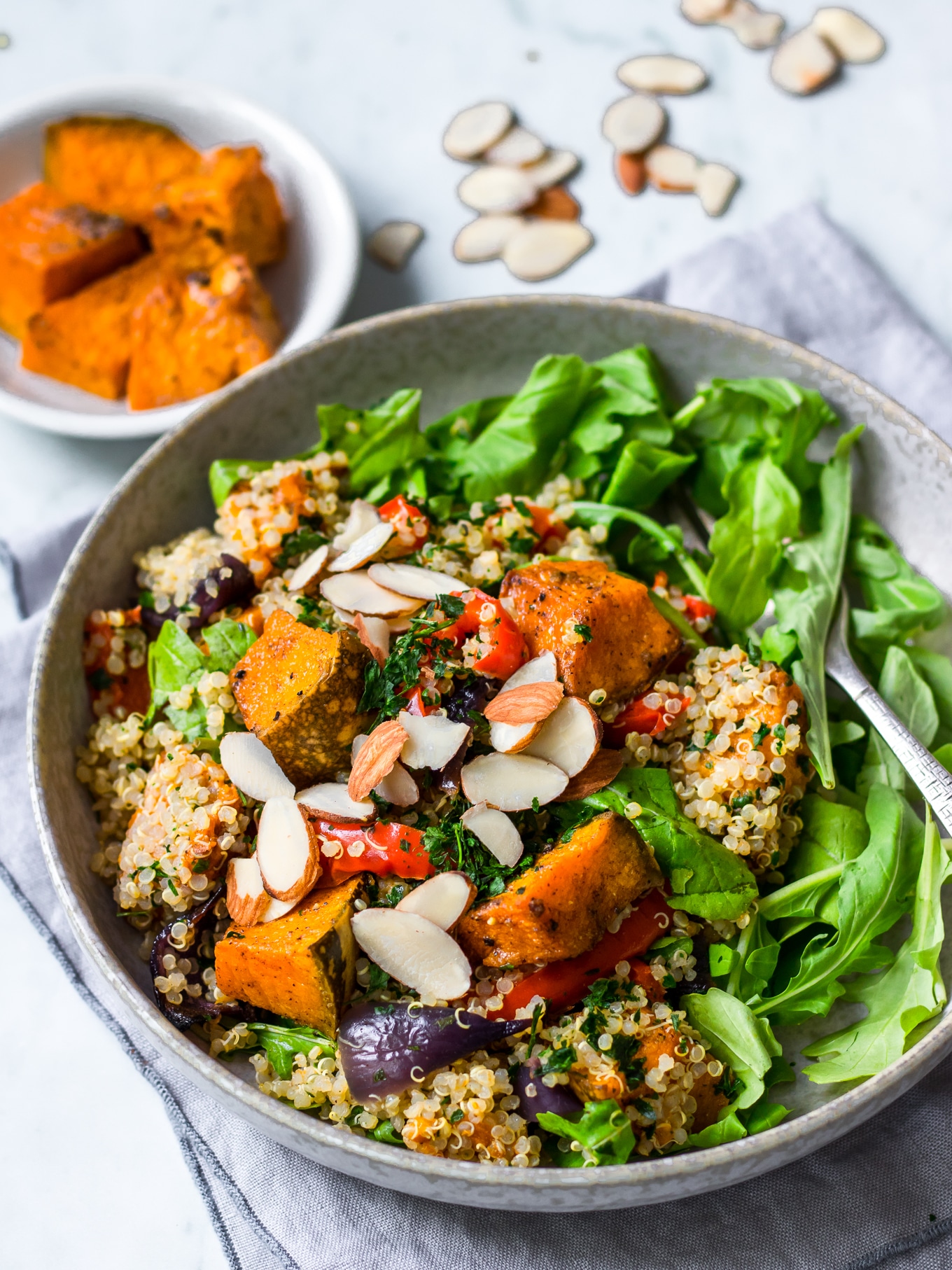 Roast pumpkin quinoa salad in grey bowl with rocket, flaked almonds on top, roast pumpkin in background