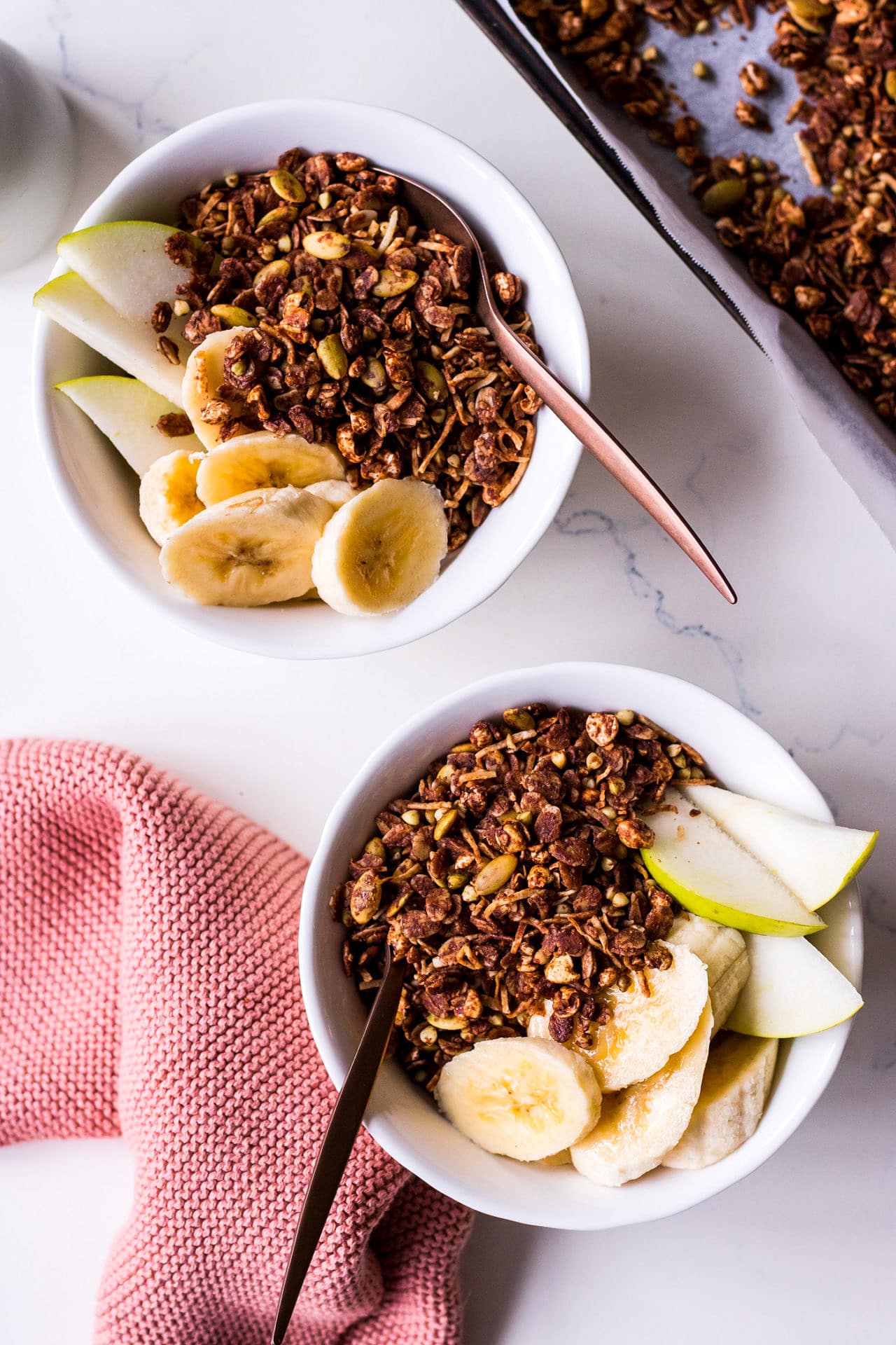 Two white cereal bowls close up spaced diagonally with banana, pear, chocolate olive oil granola and bronze spoons