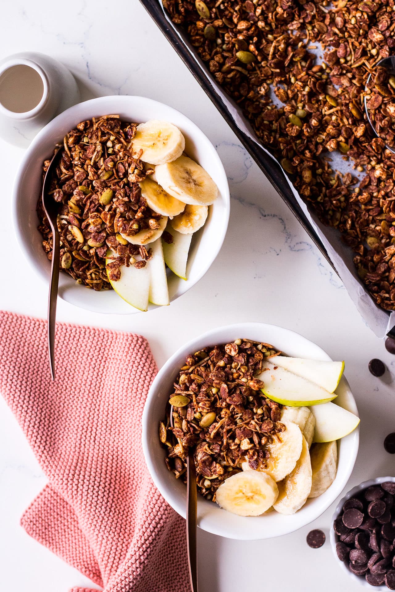 Two small white bowls placed diagonally with banana, pear, chocolate olive oil granola and bronze spoons