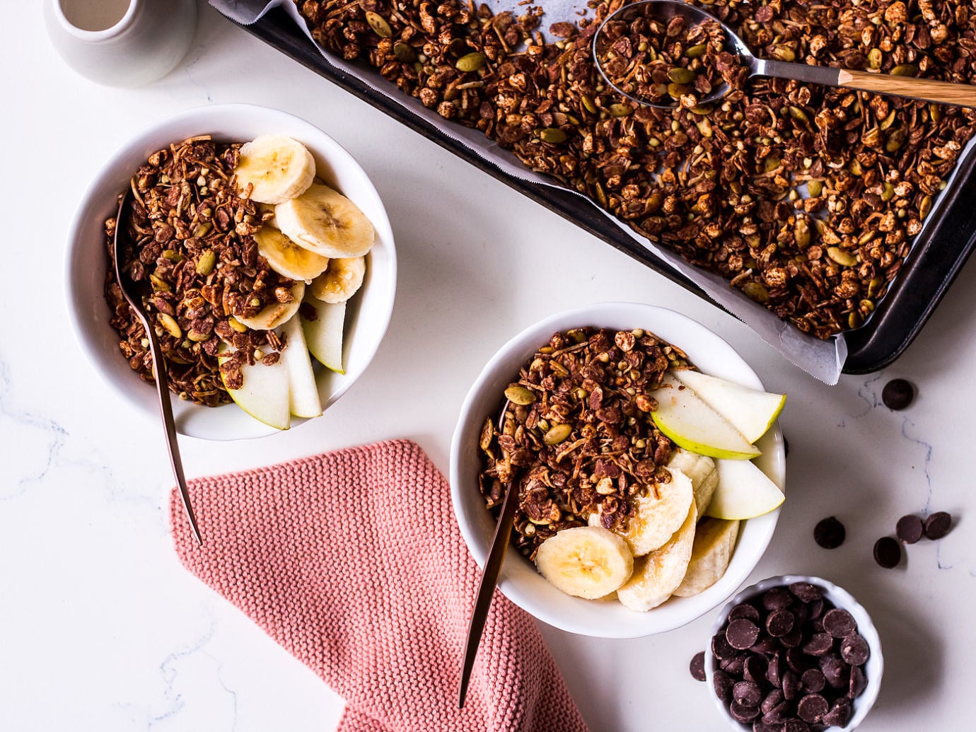 Two small white bowls with pear, banana, chocolate granola and bronze spoons, surrounded by a small cup of chocolate chips and tray of chocolate granola