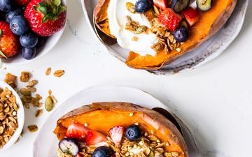 Top view of two white plates of breakfast sweet potatoes topped with yoghurt, berries and granola, with gold spoons