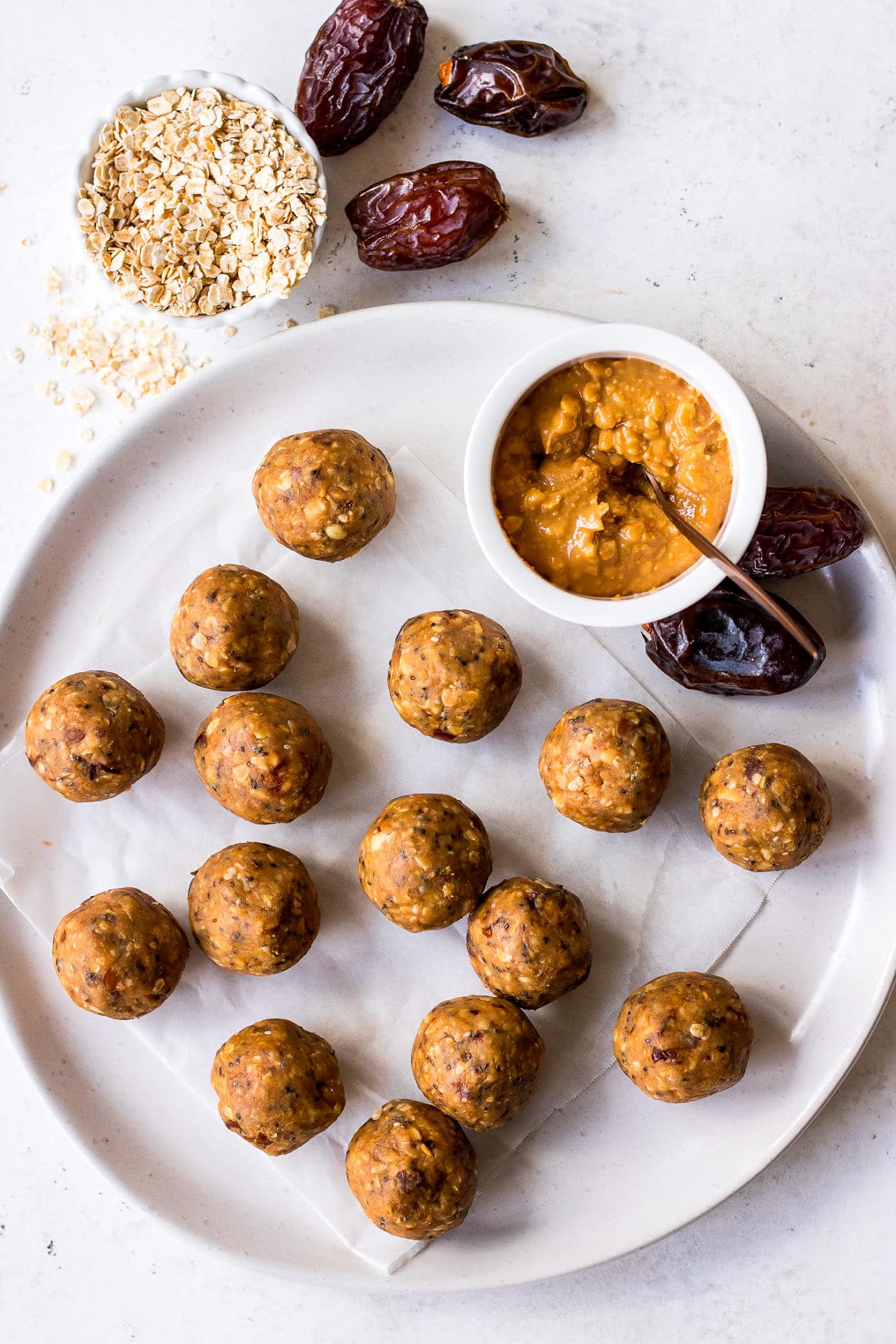 Top down view of a plate of peanut butter energy balls with a mini bowl of crunchy peanut butter, medjool dates and quick oats arranged around them