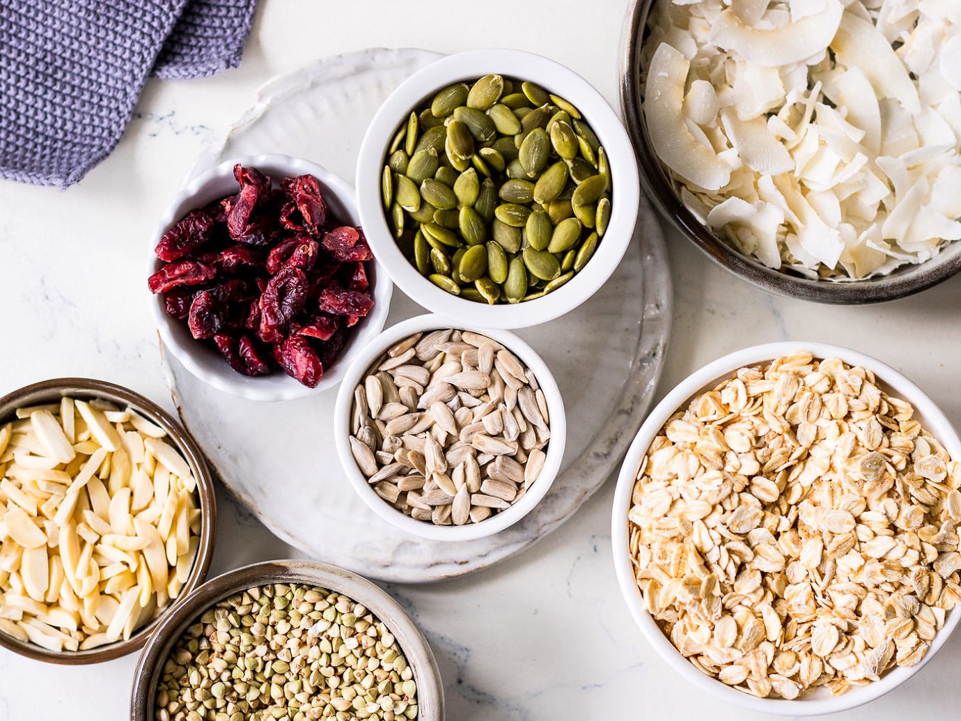White marble table with small bowls of rolled oats, coconut, pumpkin seeds, sunflower seeds cranberries, buckwheat and almonds