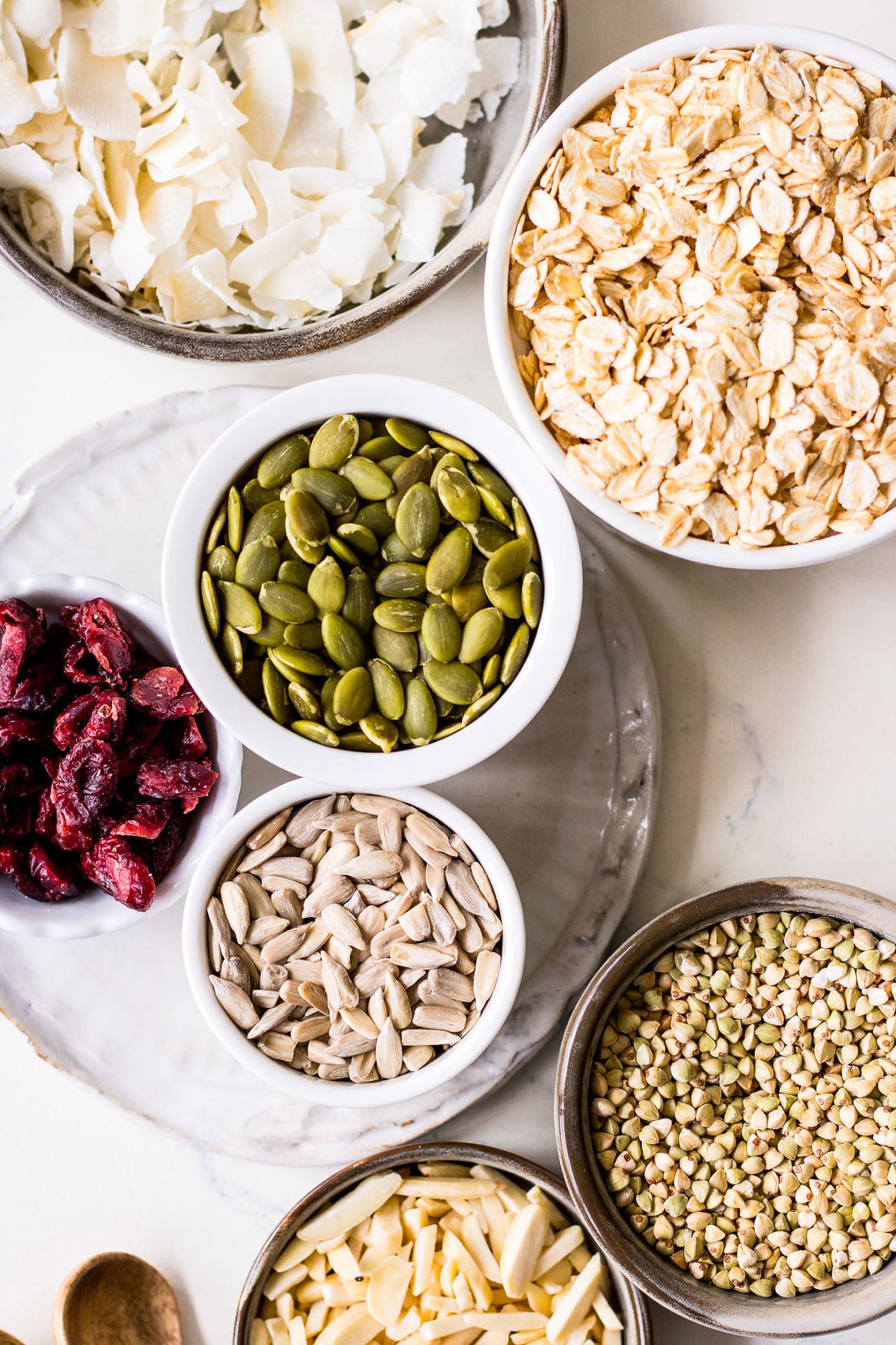 Homemade muesli ingredients arranged in small ceramic bowls. Oats, coconut flakes, cranberries, pumpkin seeds, sunflower seeds and buckwheat groats.