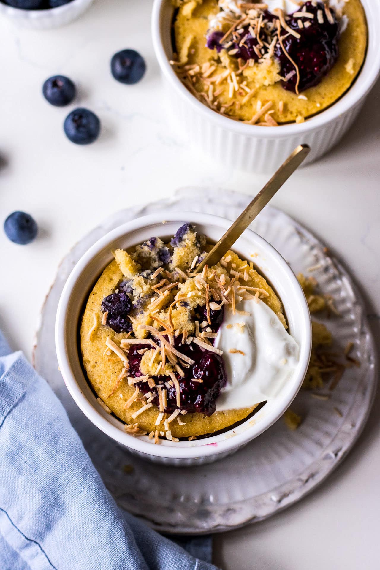Vanilla mug cake in white ramekin with blueberries, yoghurt and shredded coconut, blue napkin and blueberries