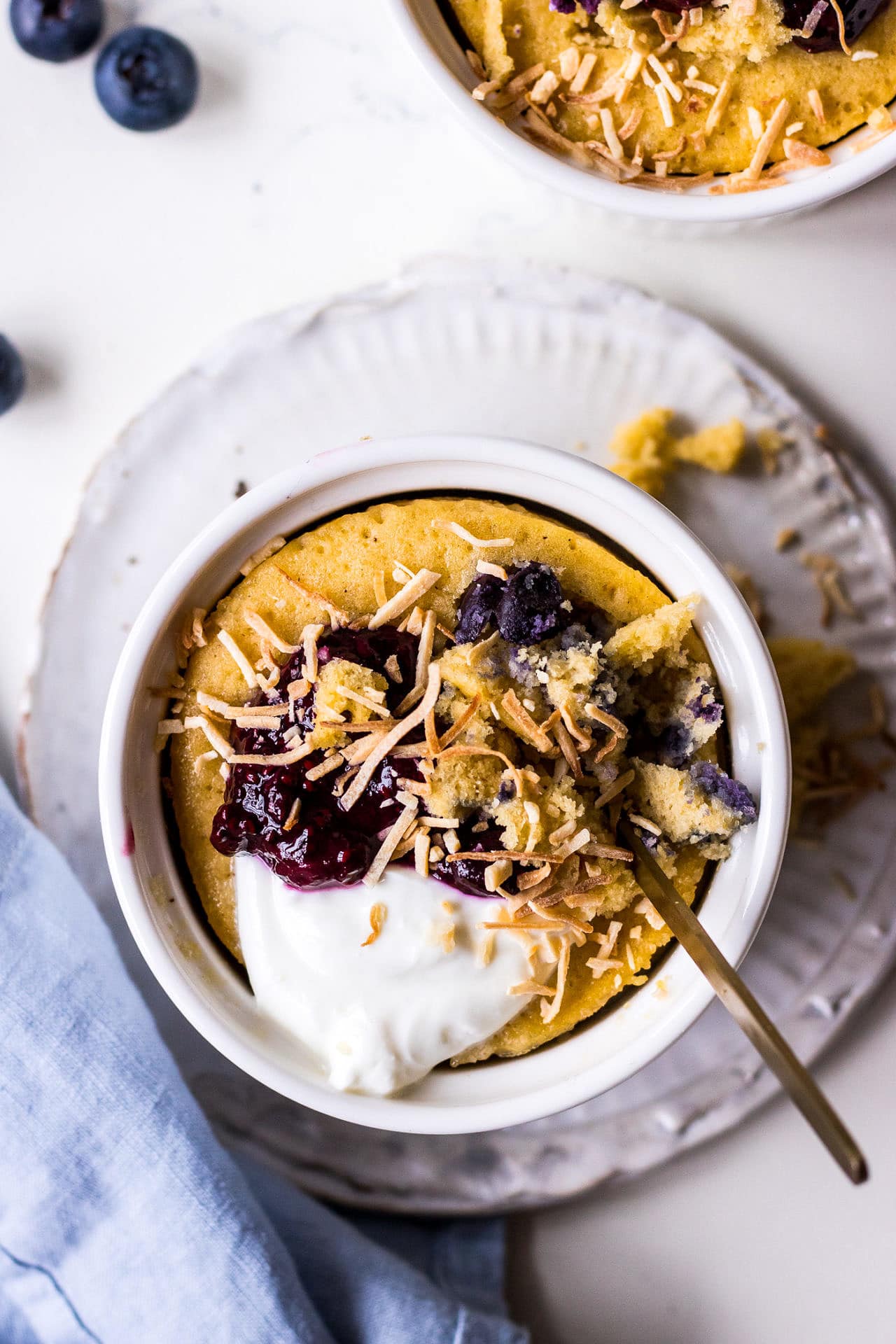 Top down view of a vanilla mug cake in white ramekin topped with blueberries, yoghurt and shredded coconut with a blue napkin and gold teaspoon