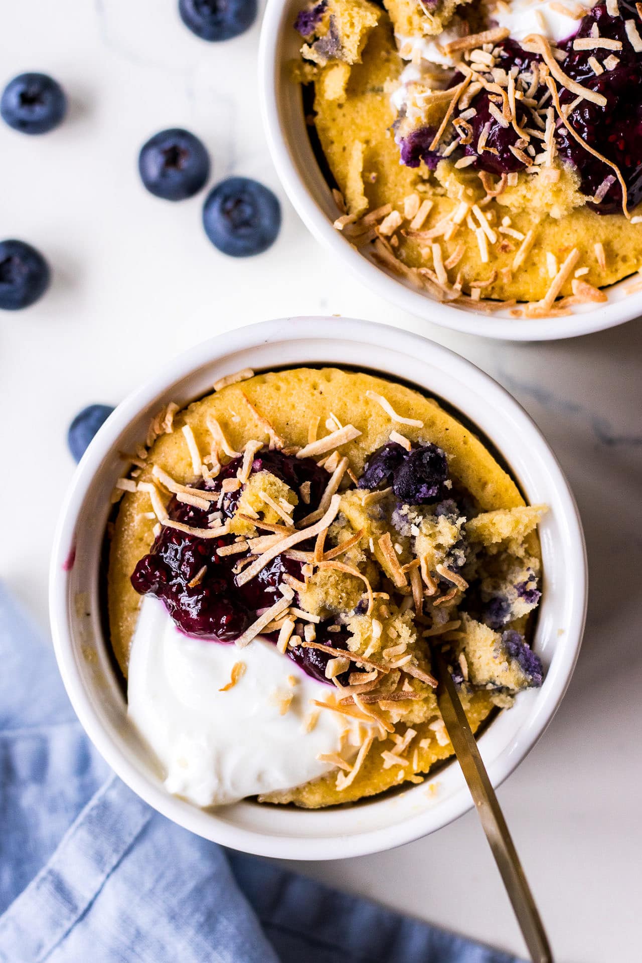 Top down view of two vanilla blueberry cakes baked in ramekins topped with yoghurt, blueberries and shredded coconut, with a blue napkin
