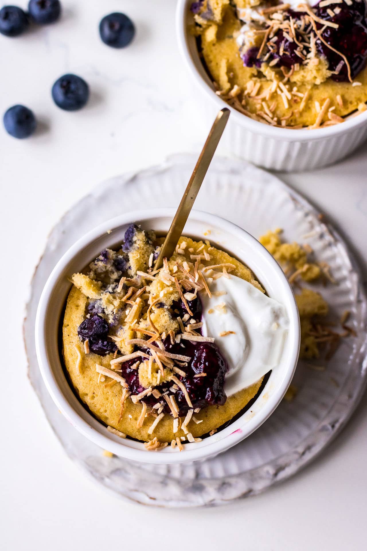 Vanilla blueberry mug cake in white ramekin on a small ceramic plate topped with blueberries, yoghurt and toasted coconut