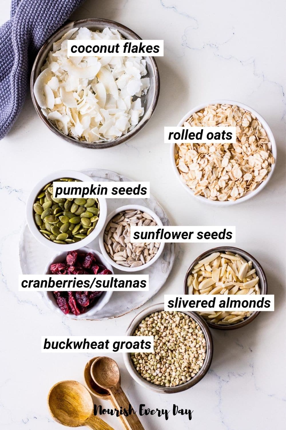Homemade muesli ingredients arranged in small ceramic bowls including rolled oats, coconut flakes, seeds, cranberries and buckwheat