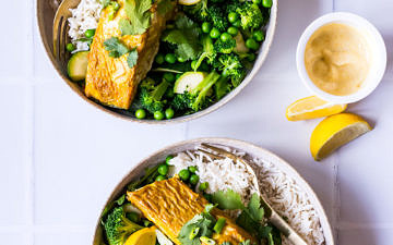 Two bowls with white rice, green vegetables and curry baked salmon garnished with lemon and coriander