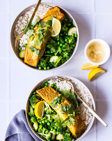 Two bowls with white rice, green vegetables and curry baked salmon garnished with lemon and coriander