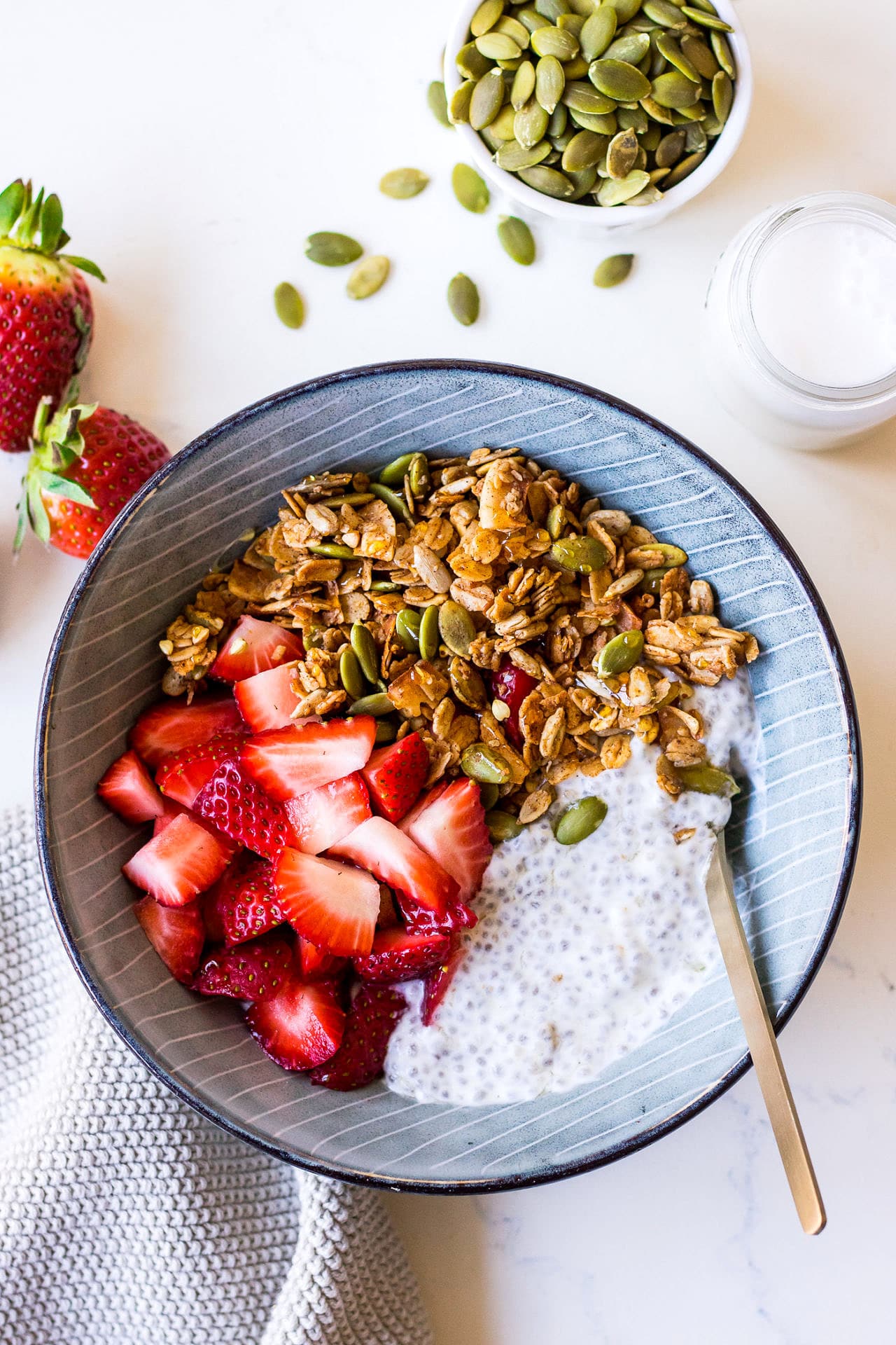 Top view blue ceramic bowl with chia pudding, chopped strawberries, granola, surrounded by a bowl of pumpkin seeds and berries