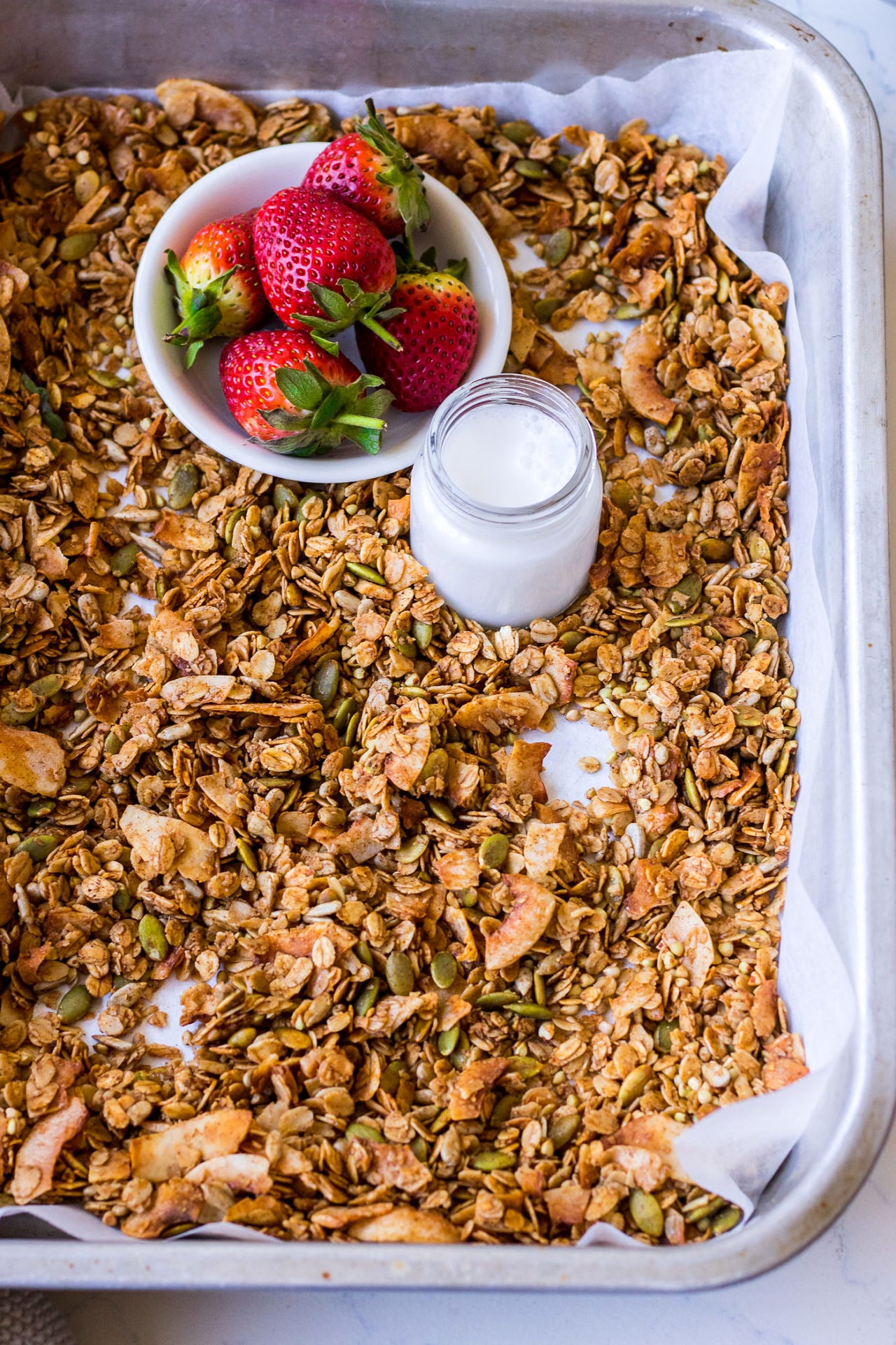 Baking tray spread out with granola, small bowl of strawberries, milk jar and small wooden spoon