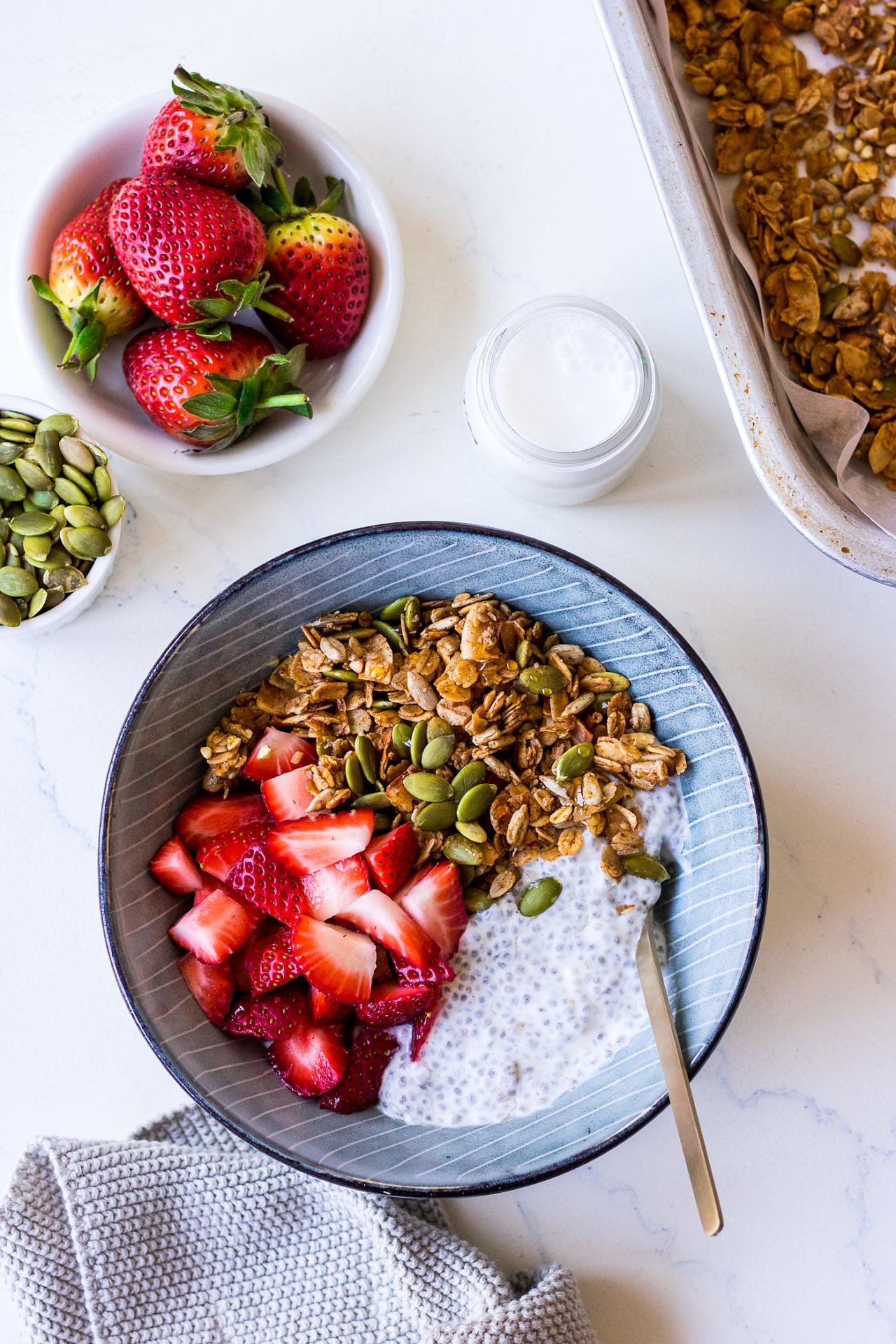 Top view blue ceramic bowl with chia pudding, chopped strawberries, granola, surrounded by a bowl of pumpkin seeds and berries