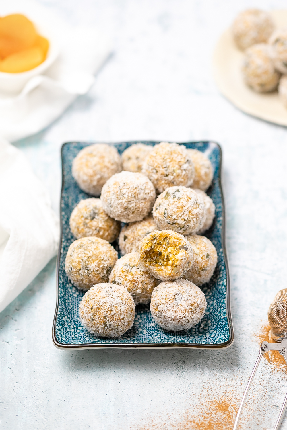 small blue rectangular plate of apricot bliss balls rolled in desiccated coconut, dish of honey in background