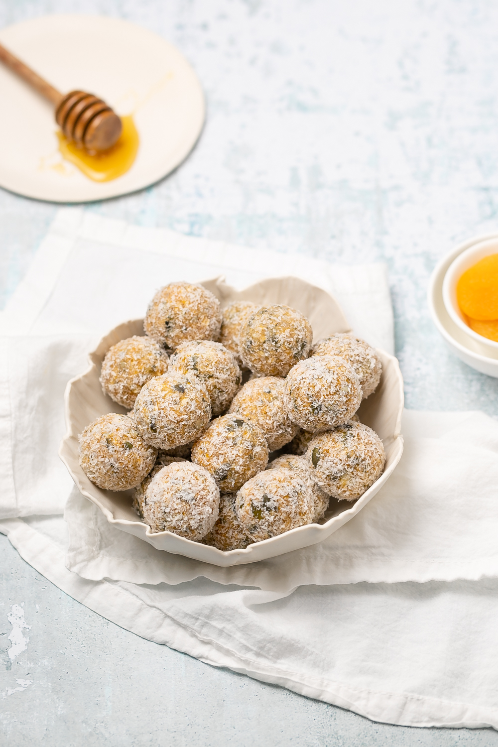 White scalloped bowl filled with apricot bliss balls coated in desiccated coconut, honey stirrer to the background, light white table