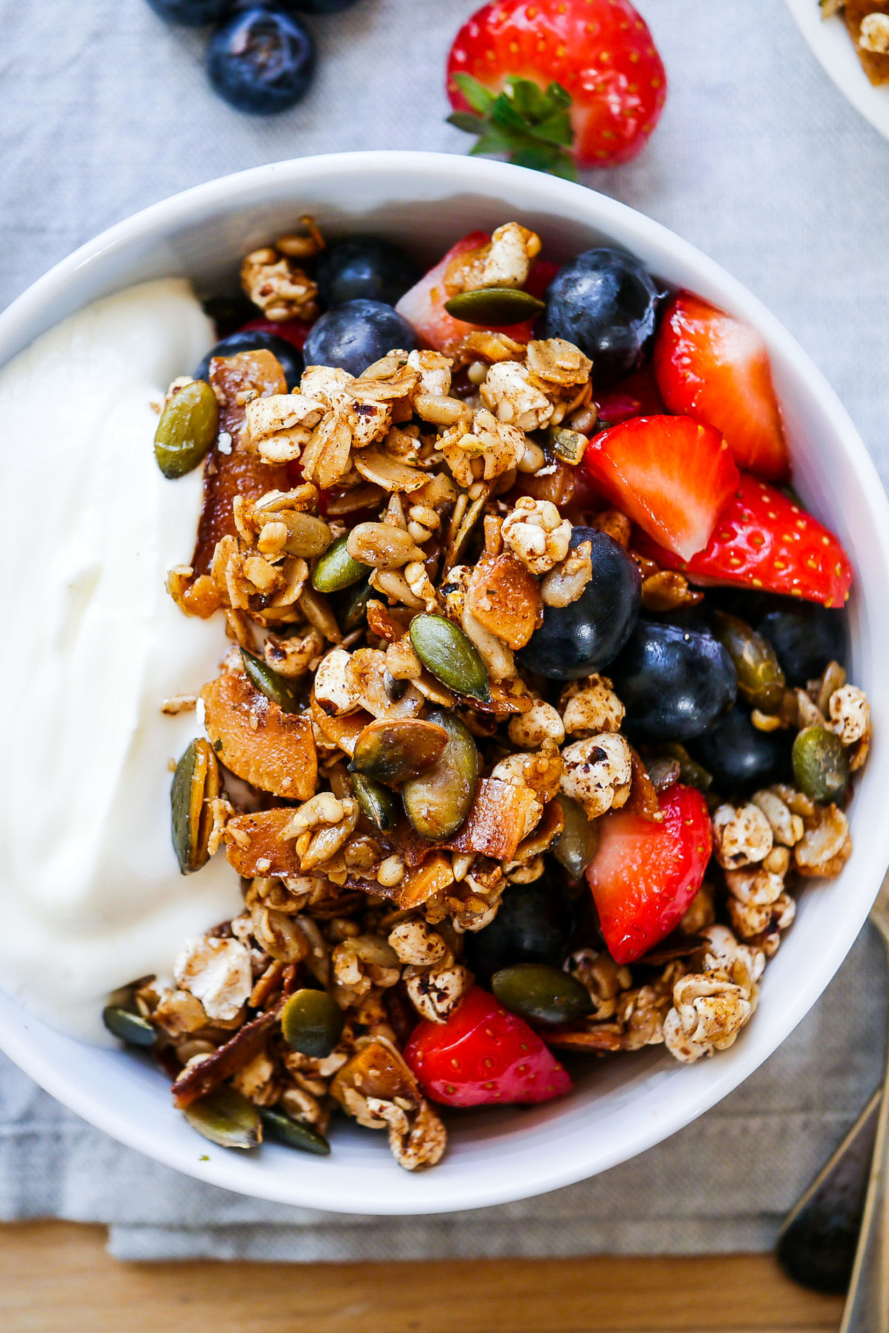 top view of pale blue cereal bowl with yoghurt, berries and toasted muesli on a wooden table, light blue napkin, vintage spoons