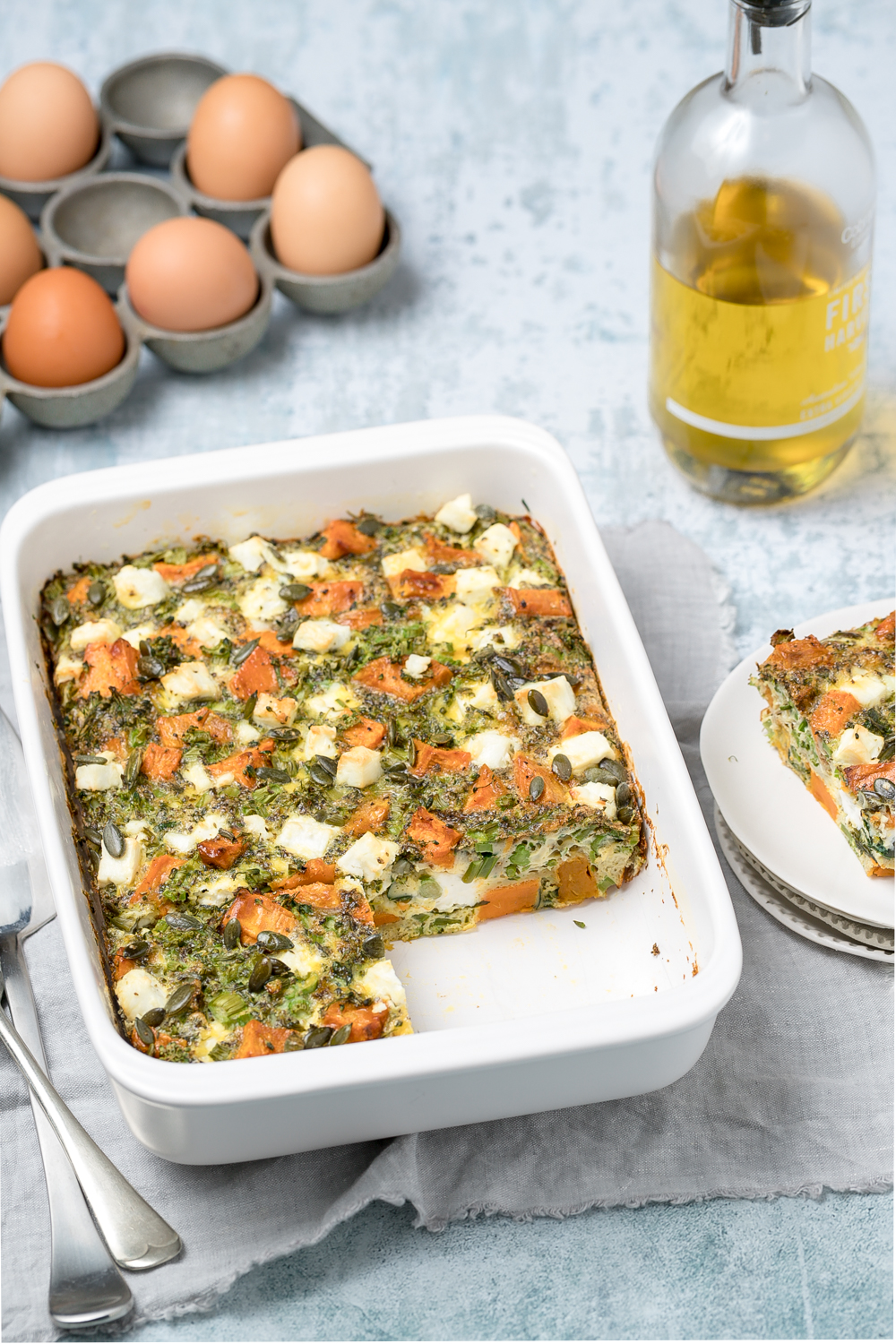 White rectangular baking dish with sweet potato, broccoli and feta frittata. Eggs and olive oil bottle in background