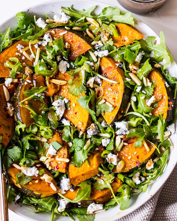 Round white ceramic dish filled with a salad of rocket, pumpkin, chilli, almonds, goats cheese and herbs