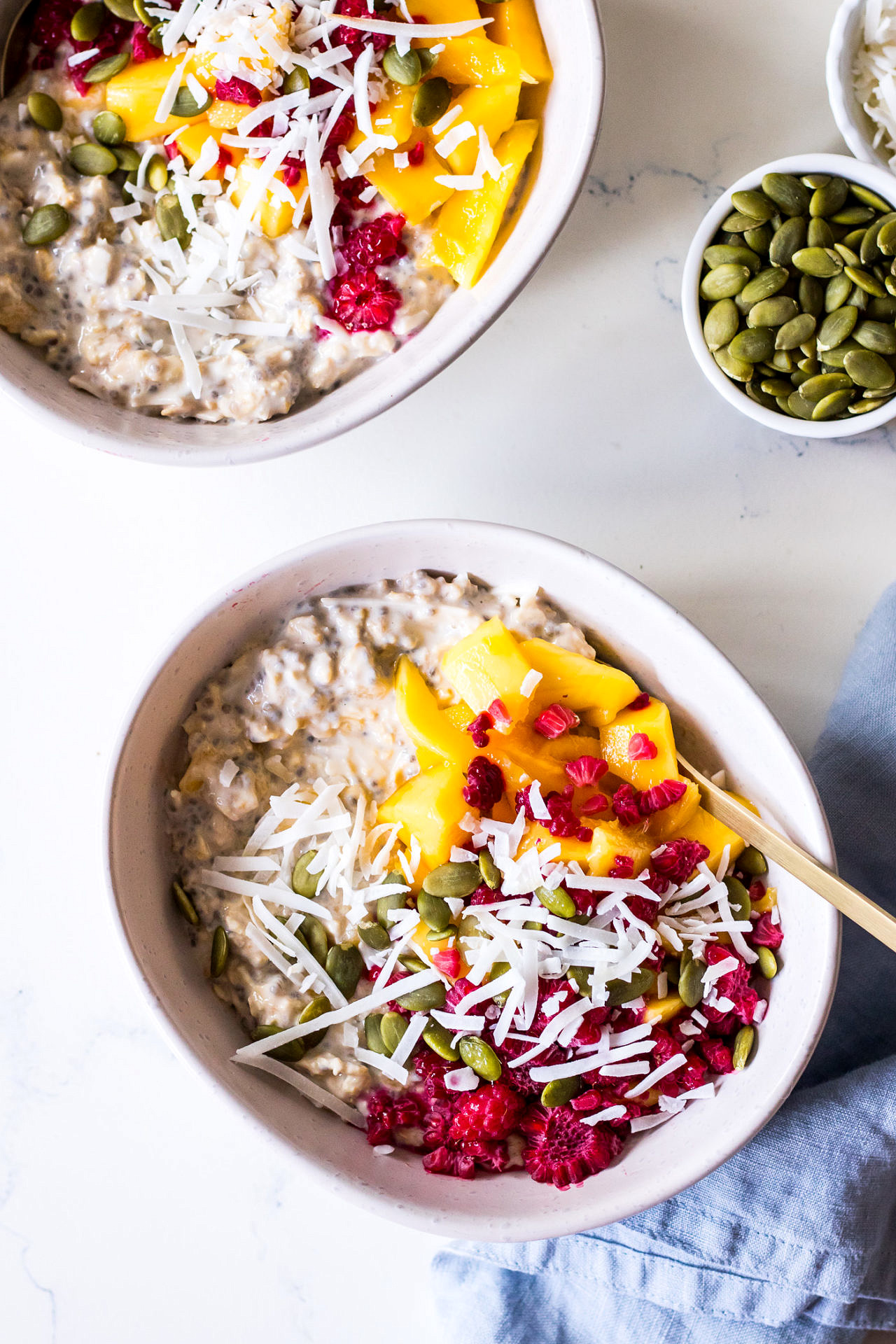 Top view of two white ceramic bowls of overnight oats with chia seeds, mango, shredded coconut and raspberries
