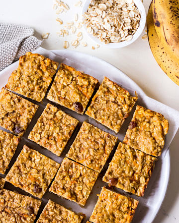 Banana oatmeal bars on a white plate with rolled oats and bananas