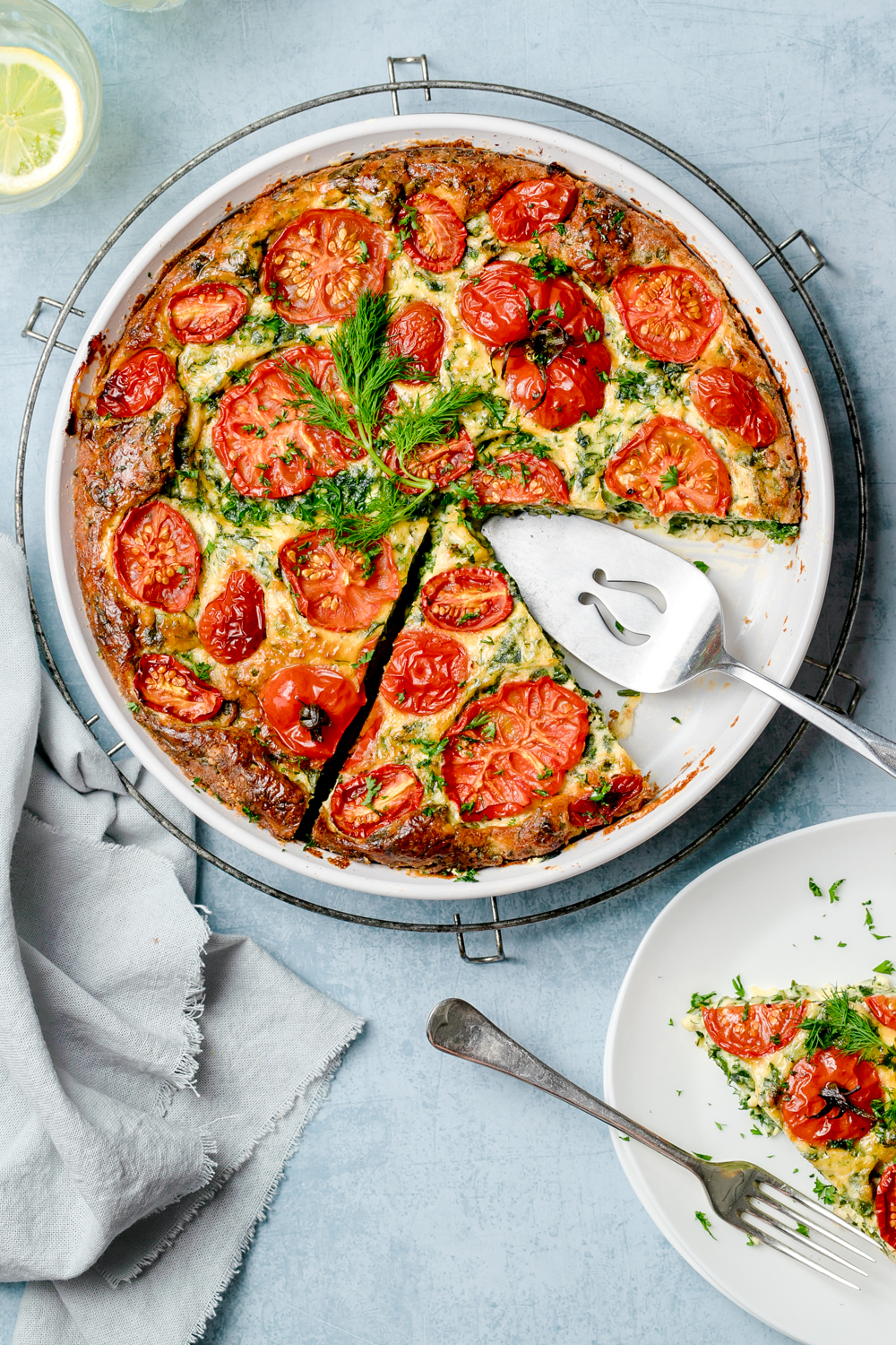 Crustless ricotta pie in a round white baking dish, made with kale and with sliced tomatoes on top. Silver serving utensil in dish.