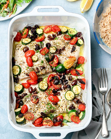 Fish tray bake with zucchini, tomato and olives on a blue background