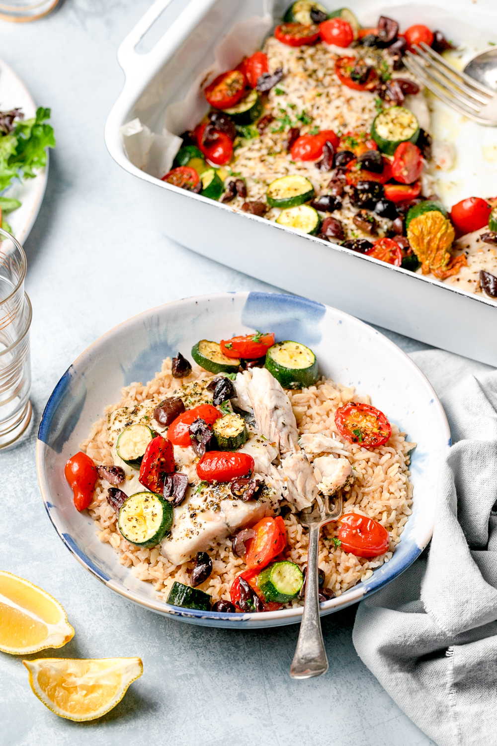 Fish, brown rice and vegetables in a shallow white bowl, fish tray bake, lemon wedges