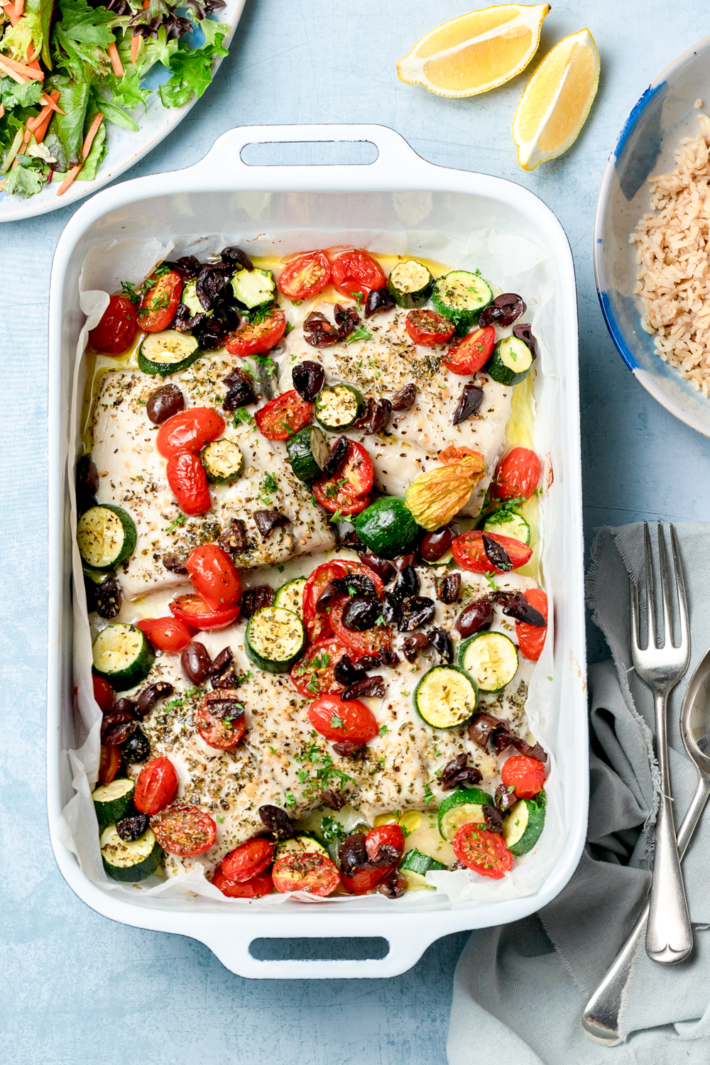Fish tray bake with zucchini, tomato and olives on a blue background