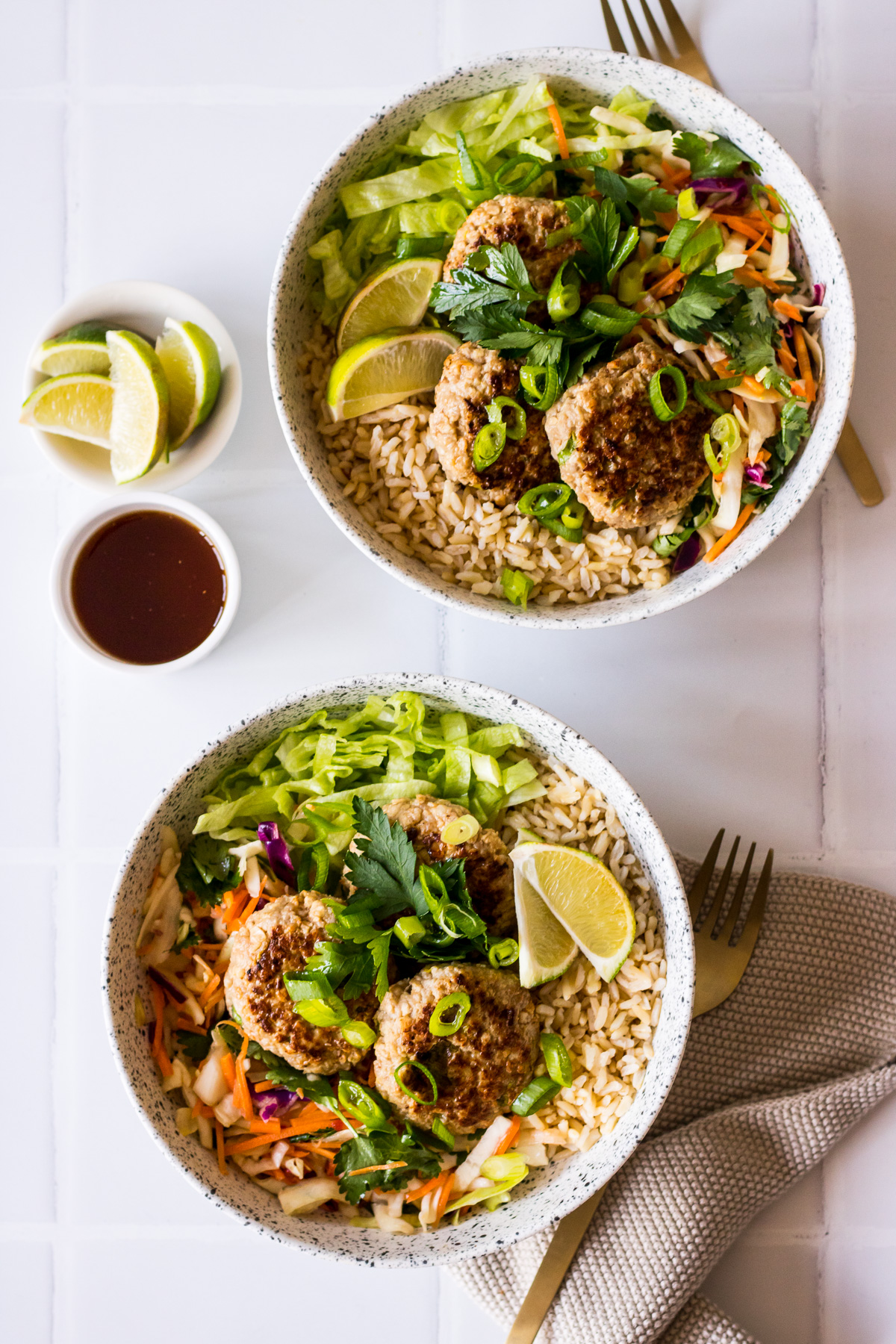 Two speckled bowls with lemongrass pork patties, brown rice, salad, soy dressing and lime wedges