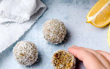 Fingers holding a lemon cashew coconut energy ball, bitten, blue table background