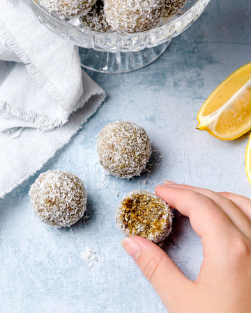 Fingers holding a lemon cashew coconut energy ball, bitten, blue table background