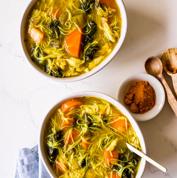 Two bowls of turmeric chicken noodle soup with gold spoons and a blue napkin