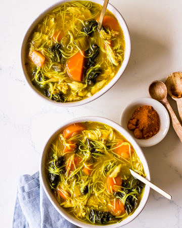 Two bowls of turmeric chicken noodle soup with gold spoons and a blue napkin