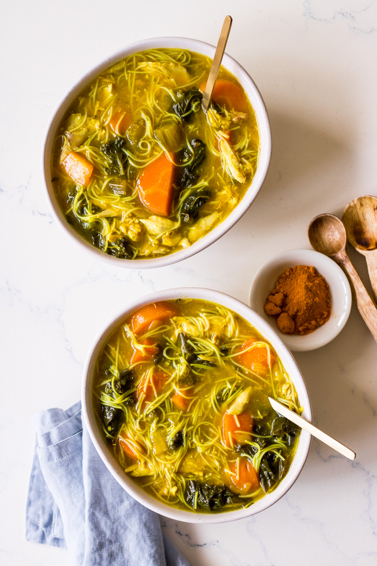 Two bowls of turmeric chicken noodle soup with gold spoons and a blue napkin
